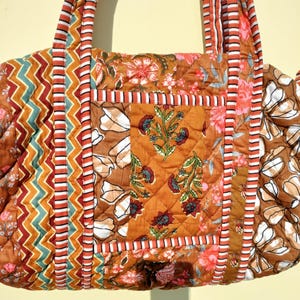 May include: A quilted tote bag with a floral pattern in shades of brown, orange, and pink. The bag features a central panel with a floral design and a diamond-stitched pattern. The handles are striped with orange, white, and brown.