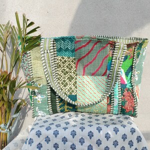 May include: A patchwork tote bag made from colorful fabric scraps. The bag has a green, blue, and white color scheme with a variety of patterns. The bag is sitting on a white fabric with a blue floral pattern.