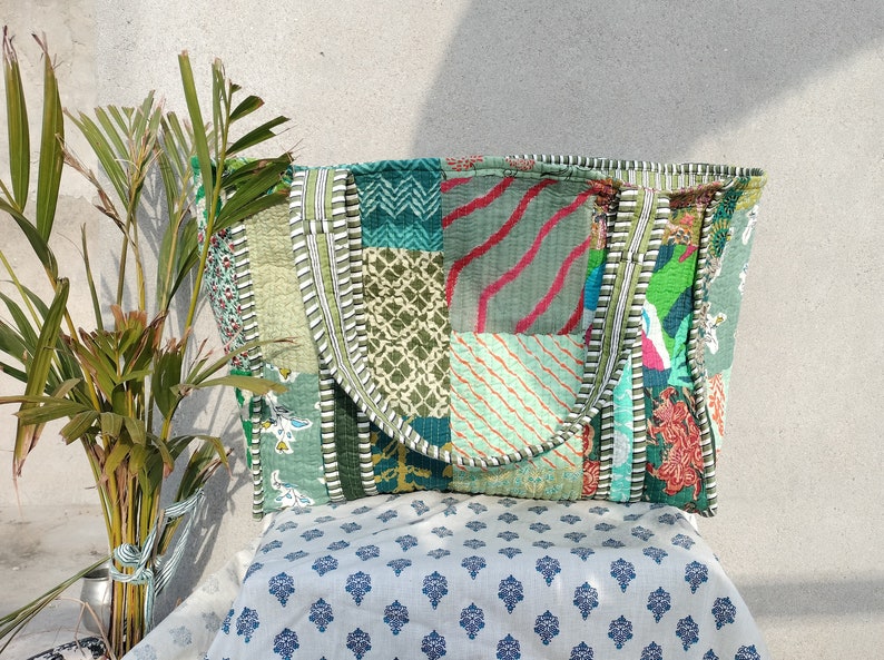 May include: A patchwork tote bag made from colorful fabric scraps. The bag has a green, blue, and white color scheme with a variety of patterns. The bag is sitting on a white fabric with a blue floral pattern.