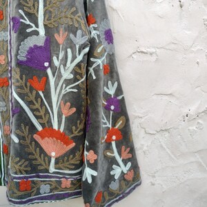 May include: Grey velvet jacket with colourful floral embroidery. The jacket features orange, purple, and peach-coloured flowers with green and white accents. The embroidery is detailed and covers the front and sleeves. It has a purple trim.