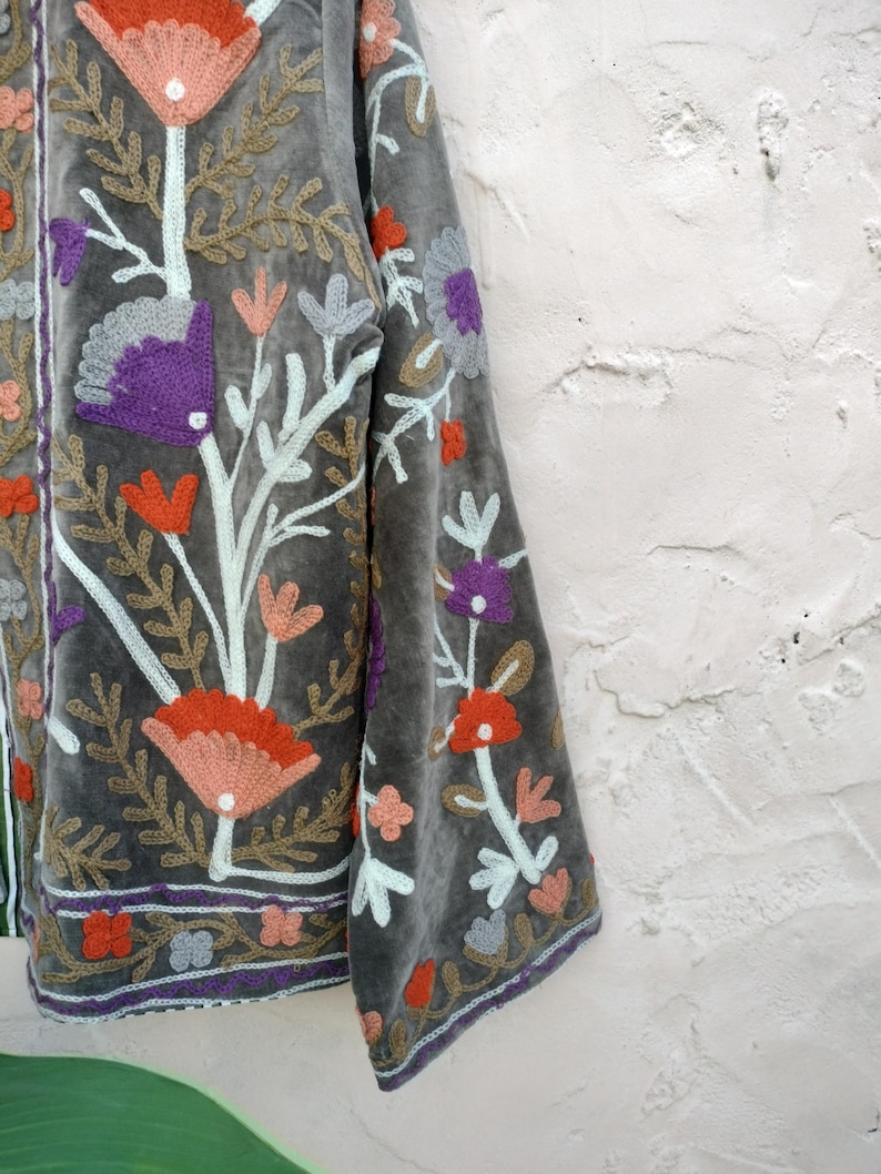May include: Grey velvet jacket with colourful floral embroidery. The jacket features orange, purple, and peach-coloured flowers with green and white accents. The embroidery is detailed and covers the front and sleeves. It has a purple trim.