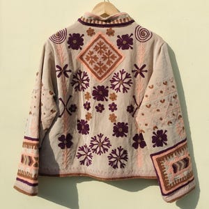 Puede incluir: Una chaqueta beige bordada con diseños florales rosas y morados. La chaqueta tiene un cuello mandarín y mangas largas con ribetes bordados.