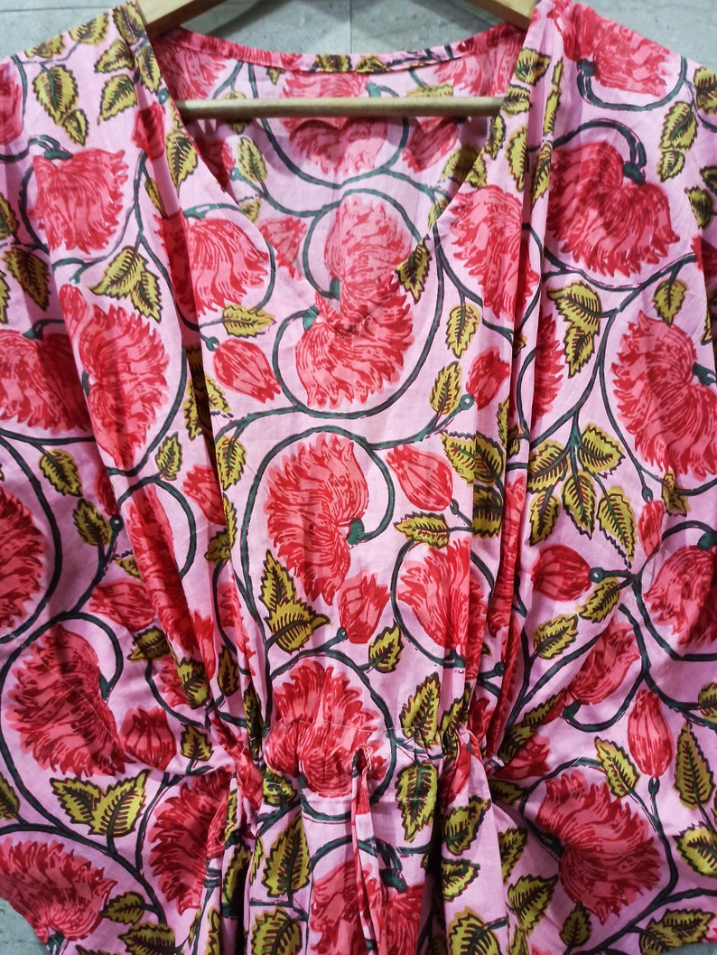 May include: A pink cotton kaftan with a floral print featuring red and green flowers. The kaftan has a V-neck and a drawstring waist.