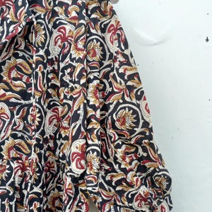 May include: A black, red, and gold floral patterned dress. The fabric features a repeating design of stylized flowers and leaves. The dress has a collared neckline and three-quarter sleeves. The garment is hanging on a wooden hanger.