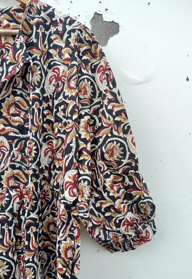 May include: A black, red, and gold floral patterned dress. The fabric features a repeating design of stylized flowers and leaves. The dress has a collared neckline and three-quarter sleeves. The garment is hanging on a wooden hanger.