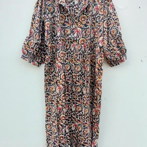 May include: A long, patterned dress with a floral design in black, red, and yellow. The dress has a collared neckline, short sleeves, and a gathered waist. It is hanging on a wooden hanger against a white wall.