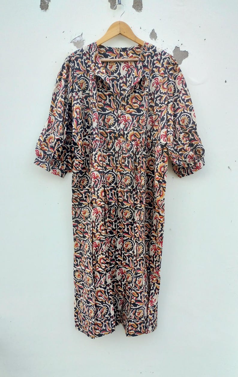 May include: A long, patterned dress with a floral design in black, red, and yellow. The dress has a collared neckline, short sleeves, and a gathered waist. It is hanging on a wooden hanger against a white wall.