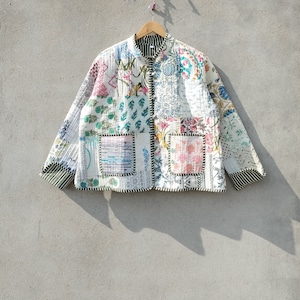 Può includere: Una giacca trapuntata bianca con un design a patchwork che presenta vari motivi floreali e geometrici in tonalità di verde, blu, rosa e bianco. La giacca ha un colletto alla coreana, una chiusura con bottone e due tasche a toppa.