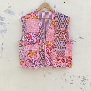 Könnte beinhalten: Eine rosafarbene gesteppte Weste mit Patchwork-Design mit floralen Mustern. Die Weste hat einen V-Ausschnitt und einen Bindeverschluss.