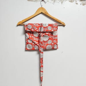 Puede incluir: Una bolsa de tela roja con un estampado floral blanco cuelga de una percha de madera. La bolsa tiene un lazo largo que se puede usar para cerrarla.