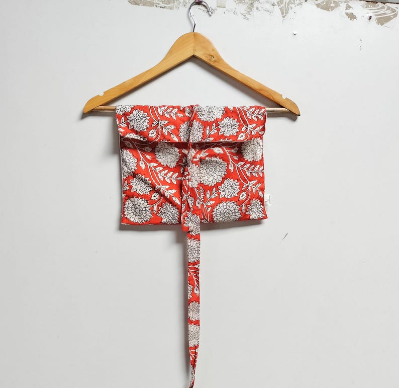 Puede incluir: Una bolsa de tela roja con un estampado floral blanco cuelga de una percha de madera. La bolsa tiene un lazo largo que se puede usar para cerrarla.