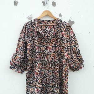 May include: A long-sleeved dress with a floral pattern in black, red, and tan. The dress is hanging on a wooden hanger against a white wall. The dress has a gathered waist and a collared neckline.