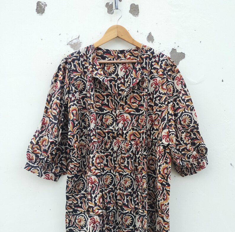 May include: A long-sleeved dress with a floral pattern in black, red, and tan. The dress is hanging on a wooden hanger against a white wall. The dress has a gathered waist and a collared neckline.
