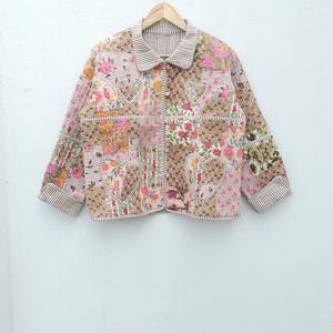 Pode incluir: Casaco acolchoado com um design de patchwork com padrões florais e em forma de coração em tons de rosa, castanho e verde. O casaco tem gola e punhos listrados e está pendurado em um cabide de madeira.