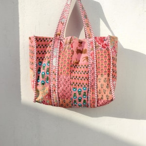 May include: A pink tote bag with floral and geometric patterns. The bag has two handles and is made of quilted fabric. The design includes shades of pink, orange, green, and blue. The bag is hanging against a white wall.