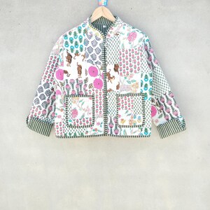 May include: A white patchwork jacket with a variety of floral and animal prints. The jacket features a mandarin collar, long sleeves, and two front pockets. The cuffs and inner collar are striped green and white. The jacket is hanging on a wooden hanger.