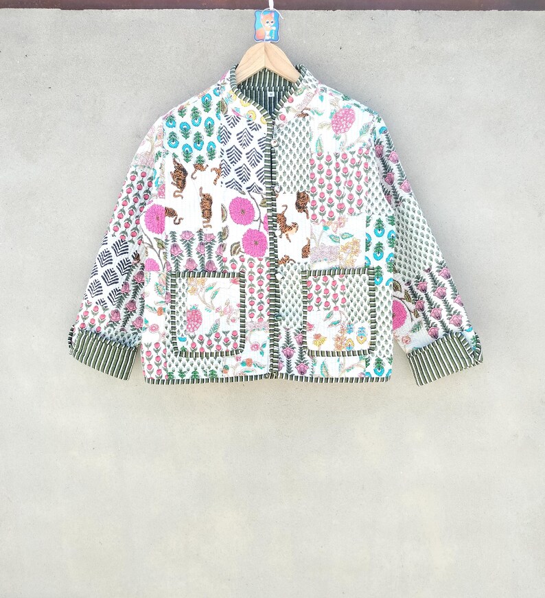 May include: A white patchwork jacket with a variety of floral and animal prints. The jacket features a mandarin collar, long sleeves, and two front pockets. The cuffs and inner collar are striped green and white. The jacket is hanging on a wooden hanger.
