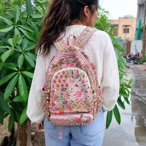 May include: A quilted backpack featuring a beige base with a multi-colored floral print in shades of pink, green, and blue. The bag includes a front zippered pocket, side pockets, adjustable shoulder straps, and white zipper trim details.