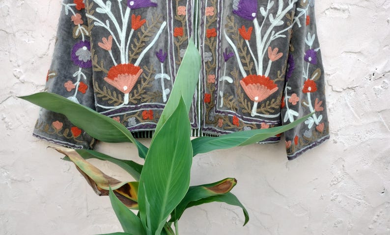 May include: A grey jacket with floral embroidery in shades of orange, purple, and pink. The jacket features a detailed design with white stems and leaves. The jacket is displayed against a white textured wall.