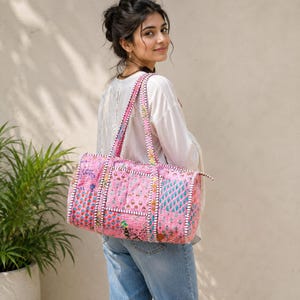 Op de afbeelding: Een cilindervormige reistas met een patchworkontwerp in roze, oranje en blauwe tinten. De tas is gemaakt van doorgestikte stof met ingewikkelde bloemen- en geometrische patronen, twee lange schouderbanden en een ritssluiting aan de bovenzijde.