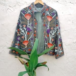 May include: A grey velvet jacket with floral embroidery in red, orange, purple, and white. The jacket has a striped lining and is hanging on a wooden hanger. A unique piece of clothing.