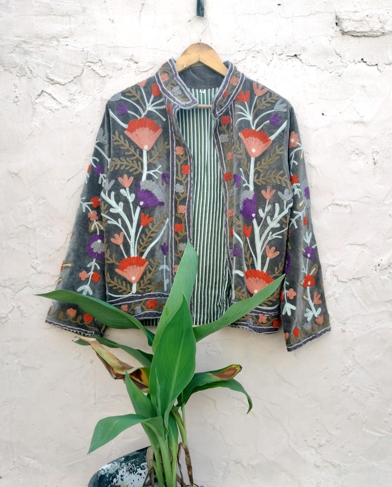 May include: A grey velvet jacket with floral embroidery in red, orange, purple, and white. The jacket has a striped lining and is hanging on a wooden hanger. A unique piece of clothing.