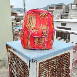 Puede incluir: Una vibrante mochila con un diseño de estampado de tigre. La mochila tiene un patrón acolchado con rayas naranjas y rosas, e ilustraciones de tigres. La bolsa tiene una parte superior redondeada, un bolsillo frontal y un ribete rosa.