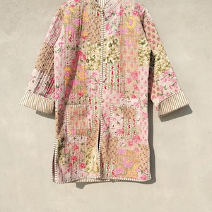 Può includere: Una giacca lunga trapuntata con un design patchwork. La giacca presenta un mix di motivi floreali e geometrici nelle tonalità del rosa, marrone e beige. Le maniche hanno polsini a righe e la giacca è appesa a una gruccia di legno.