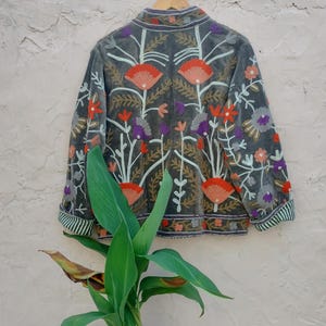 May include: A grey velvet jacket with colourful floral embroidery. The jacket features orange, purple, and white flowers, with green and white striped cuffs. The jacket is hanging on a wooden hanger against a textured wall.