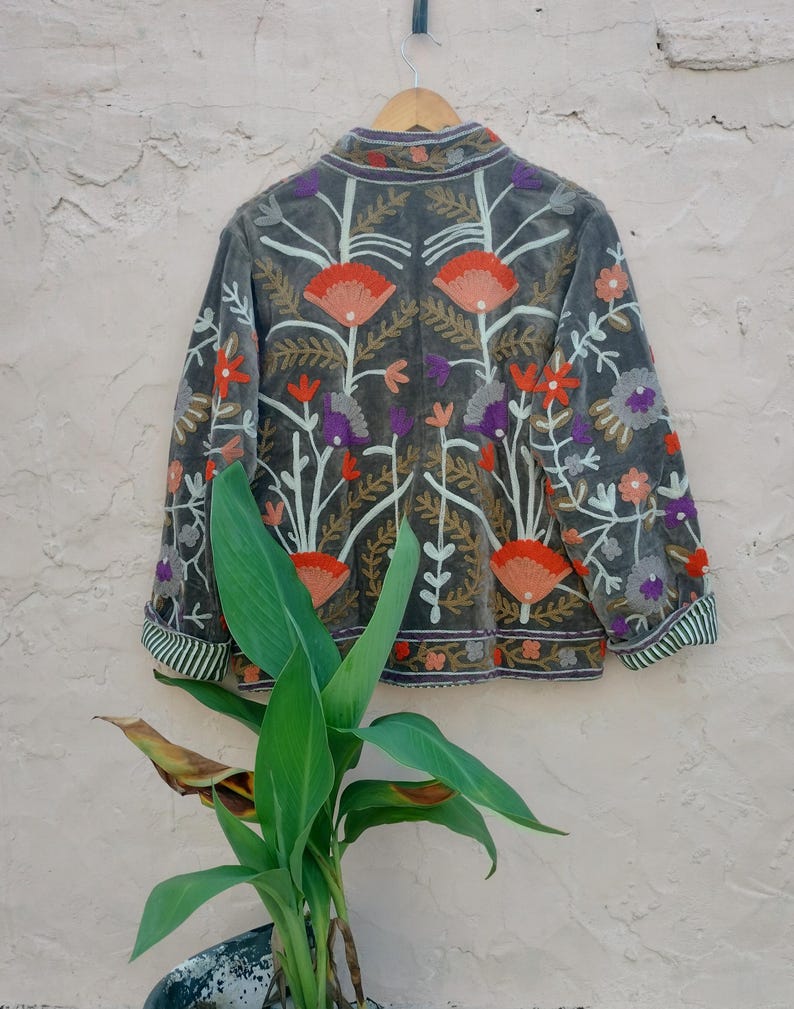 May include: A grey velvet jacket with colourful floral embroidery. The jacket features orange, purple, and white flowers, with green and white striped cuffs. The jacket is hanging on a wooden hanger against a textured wall.