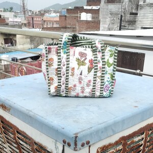 May include: A white tote bag featuring a floral design in pink, red, and green. The bag has green and white striped handles and trim. The bag is placed on a light blue surface.