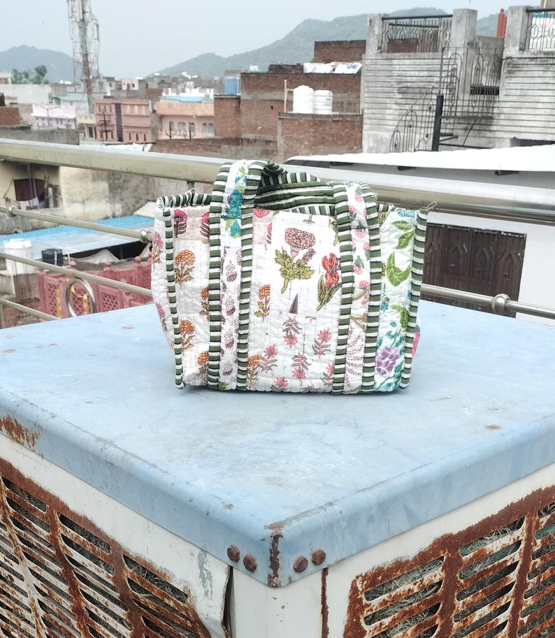 May include: A white tote bag featuring a floral design in pink, red, and green. The bag has green and white striped handles and trim. The bag is placed on a light blue surface.