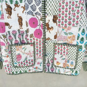 May include: A quilted jacket with a patchwork design. The jacket features various patterns, including tigers, flowers, and geometric shapes. The color palette includes white, pink, green, and brown. The jacket has two pockets and a button-up front.
