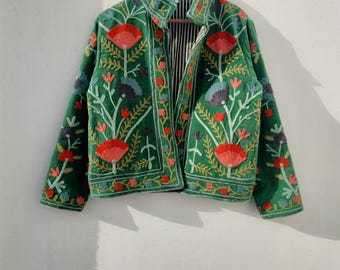 Puede incluir: Chaqueta verde con bordados florales en rojo, azul y blanco. La chaqueta tiene un cuello alto y está colgada de una percha de madera. El forro interior tiene un estampado de rayas blancas y negras.