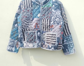 May include: A patchwork quilted jacket in blue, grey, and white. The jacket has a stand-up collar and features floral and geometric patterns. It is hanging on a wooden hanger. The jacket's design includes various textures and patterns.