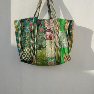 Op de afbeelding: Een patchwork tote bag met verschillende groene, turquoise en roze stofpatronen. De tas heeft twee lange handvatten en hangt tegen een witte muur. Het ontwerp bevat bloemen-, geometrische en gestreepte patronen.