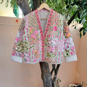 May include: A beige floral-embroidered jacket with pink, yellow, and green accents. The jacket features a quilted design, two pockets, and striped cuffs. It is displayed on a wooden hanger, set against a natural backdrop.