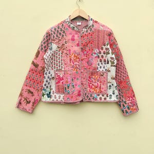 May include: A quilted jacket with a patchwork design in shades of pink, red, and white. The jacket features a mandarin collar and is hung on a wooden hanger. The fabric is patterned with floral and geometric motifs.