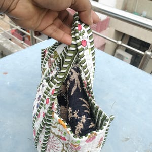May include: A quilted fabric bag with a floral pattern in pink, green, and white. The bag has a striped green and white border and contains a rolled-up black fabric with a floral design. The bag is held open.