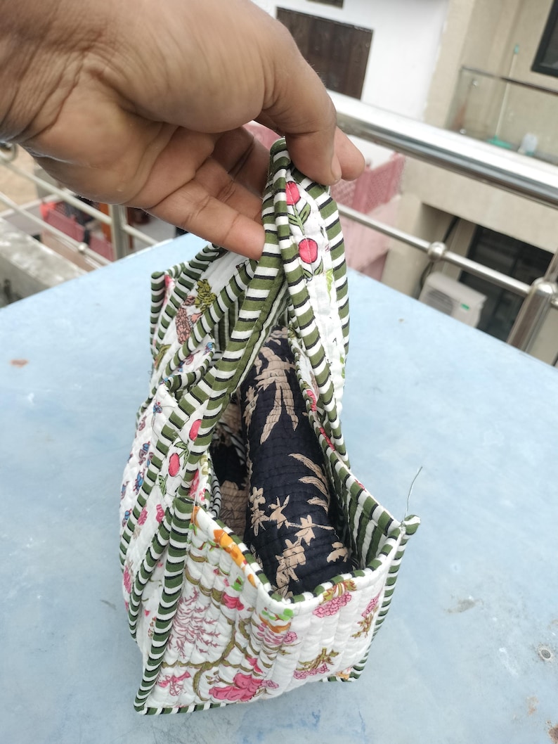 May include: A quilted fabric bag with a floral pattern in pink, green, and white. The bag has a striped green and white border and contains a rolled-up black fabric with a floral design. The bag is held open.
