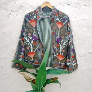 May include: A grey velvet jacket with floral embroidery in red, orange, purple, and white. The jacket has a striped lining and is hanging on a wooden hanger against a textured wall. The jacket's design features a collar and long sleeves.