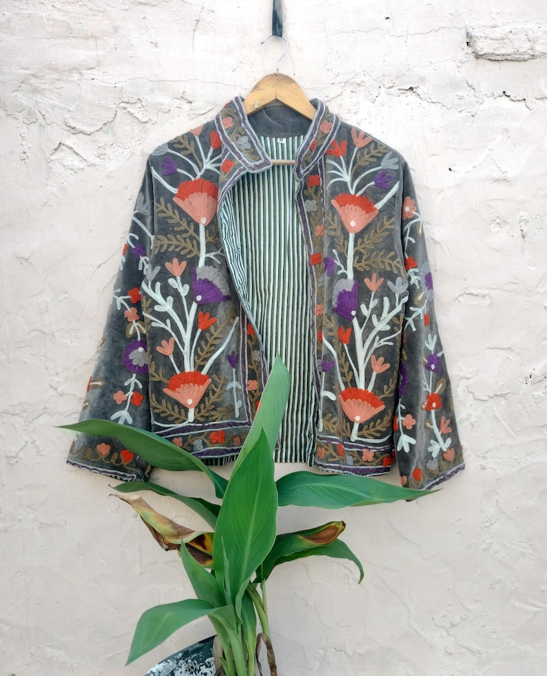 May include: A grey velvet jacket with floral embroidery in red, orange, purple, and white. The jacket has a striped lining and is hanging on a wooden hanger against a textured wall. The jacket's design features a collar and long sleeves.