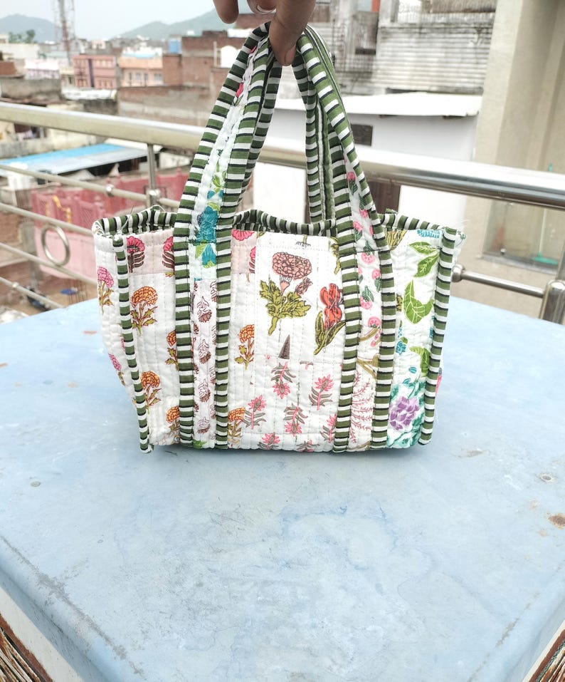 May include: A white quilted tote bag featuring a floral design in pink, green, and orange. The bag has green and white striped handles and trim. The bag is approximately 30 cm wide and 25 cm tall.