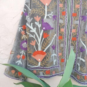 May include: A grey velvet jacket with detailed floral embroidery in orange, purple, and gold. The jacket has an open design with elaborate patterns on the edges and sleeves, offering a bohemian style.
