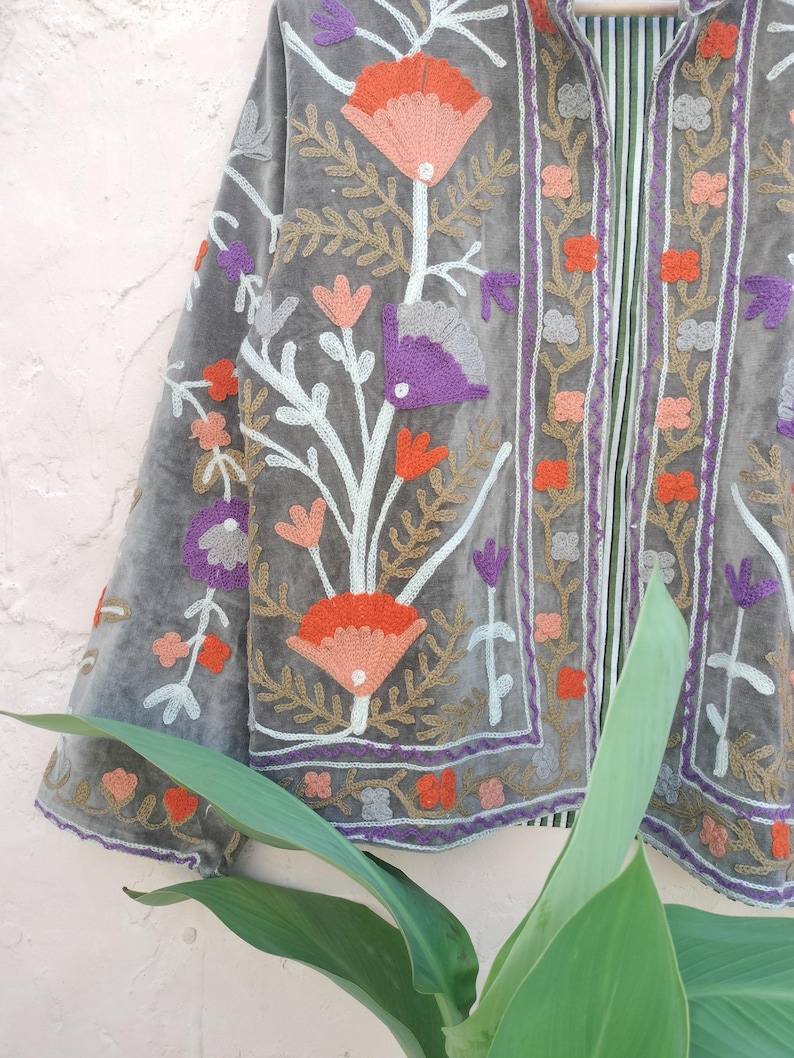 May include: A grey velvet jacket with detailed floral embroidery in orange, purple, and gold. The jacket has an open design with elaborate patterns on the edges and sleeves, offering a bohemian style.