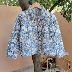 May include: A quilted jacket with a floral pattern in shades of blue, gray, and white. The jacket has a striped collar and two front pockets. It is hanging on a wooden hanger.