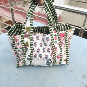 May include: A quilted tote bag featuring floral and botanical patterns in pink, blue, and green on a white backdrop. The bag has green and white striped handles and trim. The bag is held up by a hand.
