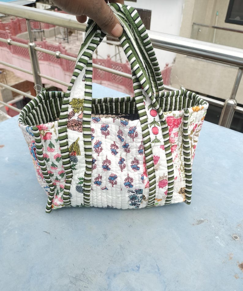 May include: A quilted tote bag featuring floral and botanical patterns in pink, blue, and green on a white backdrop. The bag has green and white striped handles and trim. The bag is held up by a hand.