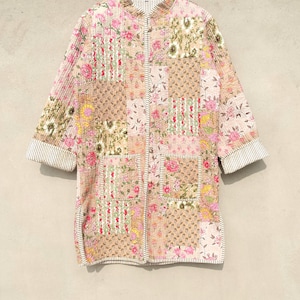 May include: A patchwork jacket with a floral pattern in pink, beige, and green. The jacket has a mandarin collar, two pockets, and three-quarter sleeves with striped cuffs. The jacket is hanging on a wooden hanger.