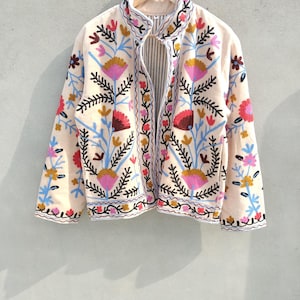 Peut inclure: Veste crème avec broderies florales vibrantes. La veste a un col montant et des manches longues. La broderie comprend des fleurs roses, rouges, bleues et jaunes avec des accents de feuilles noires. La veste est suspendue à un cintre en bois.